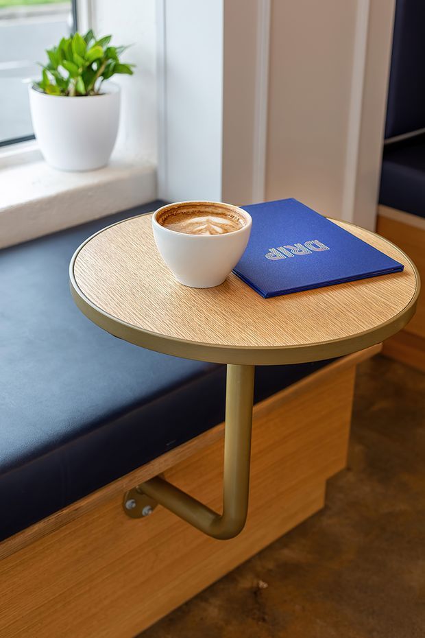 Close-up interior detail of a compact built-in window seat with dark upholstered cushioning and timber cabinetry beneath. A round light timber side table with a brass-toned edge and tubular support holds a coffee cup and blue booklet. A potted plant sits on the deep window sill, with warm timber and dark flooring contributing to the setting.