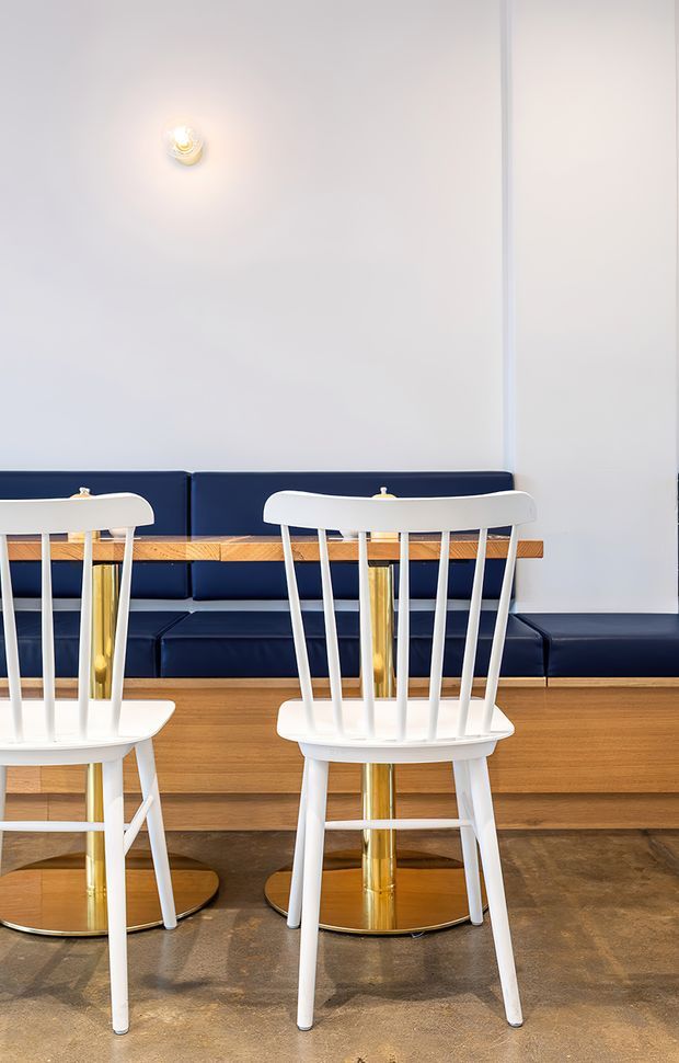 A refined dining or café-style seating area with two white spindle-back chairs in the foreground, each supported by polished brass pedestal bases. A long dark navy upholstered banquette runs along the wall behind a narrow timber ledge, with a simple wall sconce above. The space combines pale walls, warm timber joinery, navy upholstery, brass accents, and a polished concrete floor in a clean, contemporary composition.
