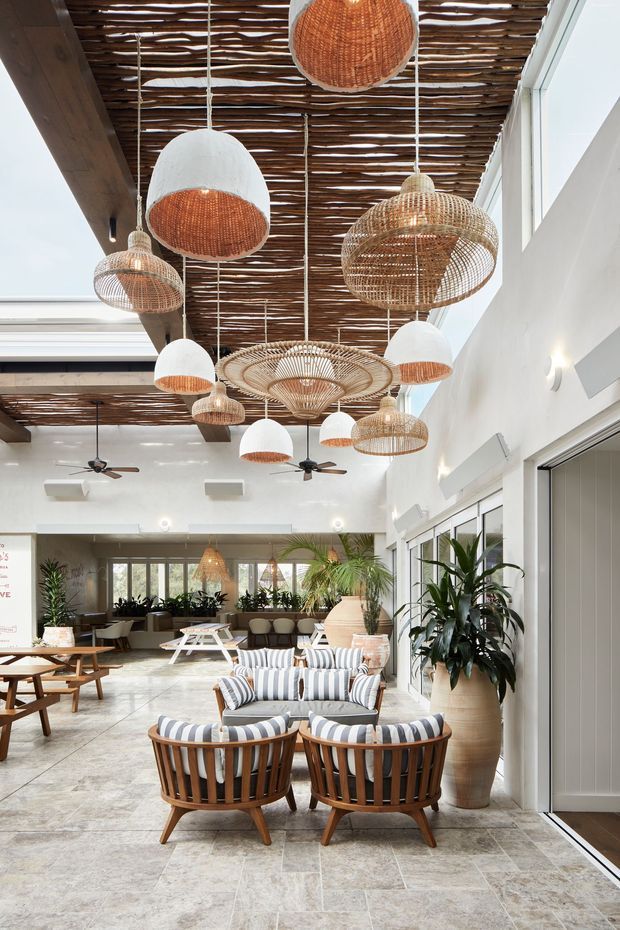 Bright, spacious indoor lounge or hospitality space with white walls, large glazing, timber beams, and a striking ceiling installation of woven rattan pendant lights beneath a slatted timber canopy. The room contains striped upholstered lounge chairs and sofas, timber tables, large planters, ceiling fans, and pale stone-look tiled flooring. The overall atmosphere is airy, relaxed, and coastal-inspired.