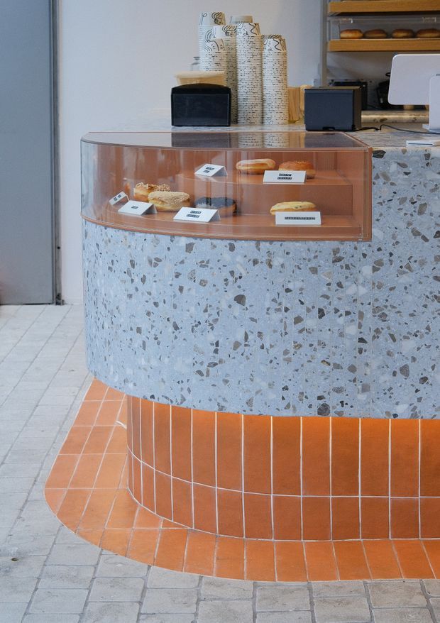 A close-up view of a curved bakery or café service counter. The lower counter is clad in speckled terrazzo, with a rounded display case above containing pastries and labels. The counter base is finished in glossy terracotta-orange square tiles with pale grout, while bread shelves, small appliances, and paper packaging are visible behind it. The space has a contemporary, tactile material palette with warm orange and cool grey tones.