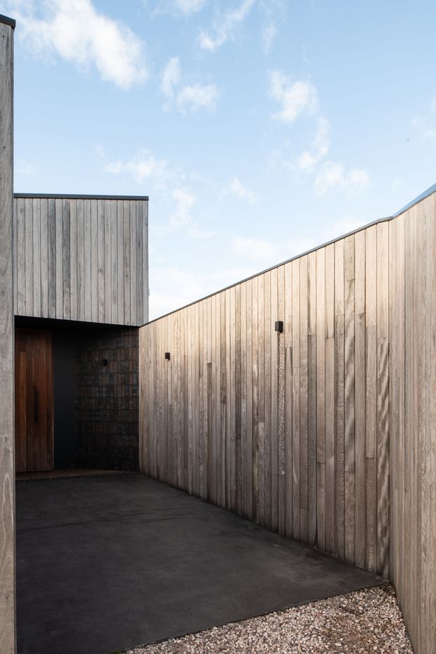 A minimalist exterior courtyard or entry approach enclosed by tall vertical timber-clad walls. The space has a dark concrete paving surface, a recessed dark timber door, black trim and wall-mounted lights, with a gravel strip in the foreground. The composition emphasizes geometric rooflines, textured weathered wood, and open sky.