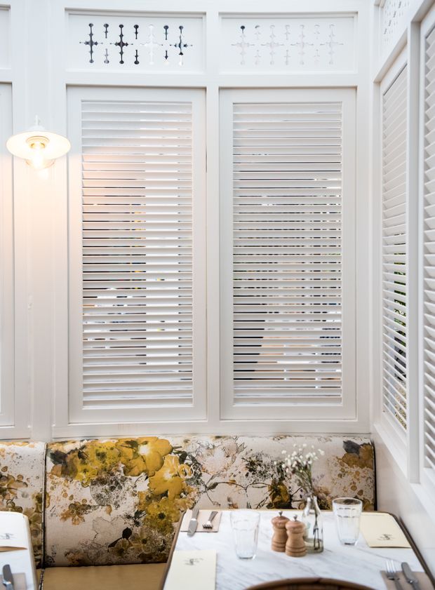 Bright, compact dining nook with white plantation shutters covering multiple windows, decorative upper panels, and white trim. A floral upholstered banquette surrounds a marble-topped dining table set with glassware and tableware. A wall-mounted light adds warm illumination.