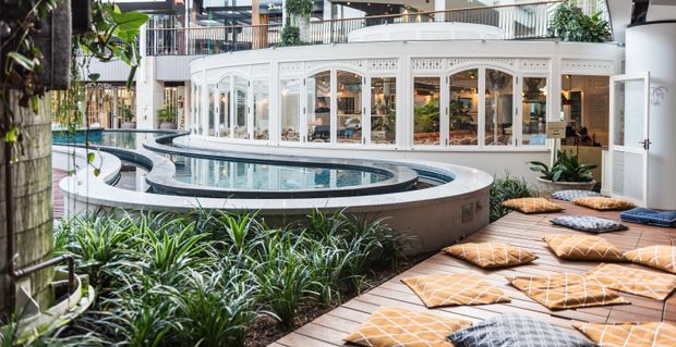 A lush indoor atrium featuring a curved raised swimming pool or water feature, surrounded by tropical planting and a timber deck with patterned floor cushions. A bright white glazed pavilion with arched windows and doors forms the backdrop, with multi-level commercial or hospitality-style interiors visible beyond.