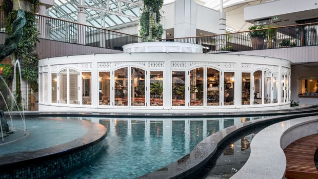 Interior atrium of a large contemporary commercial or hospitality building, centred on a curved indoor pool with tiered edges. A white, classically detailed glazed pavilion or dining enclosure sits beside the water, with upper-level balconies, lush integrated planting, and a large glass roof creating a bright conservatory-like environment.
