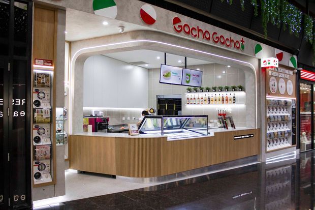 A modern shopping-centre food and beverage storefront for Gocho Gochot+, featuring a curved illuminated white service opening, warm wood-look counter cabinetry, refrigerated display cases, tiled back wall, menu screens, product shelving, and polished dark flooring.