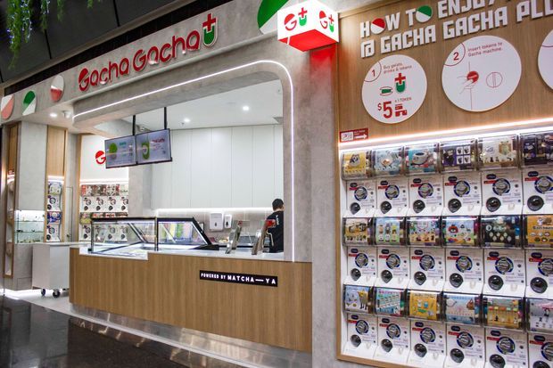 A brightly lit Gocha Gocha matcha and dessert retail kiosk in a shopping-centre setting. The compact service counter has light wood-look fixed cabinetry, glass display cases, menu screens, white wall panels, and prominent illuminated branding. A large wall-mounted retail display on the right presents packaged products and instructional graphics.