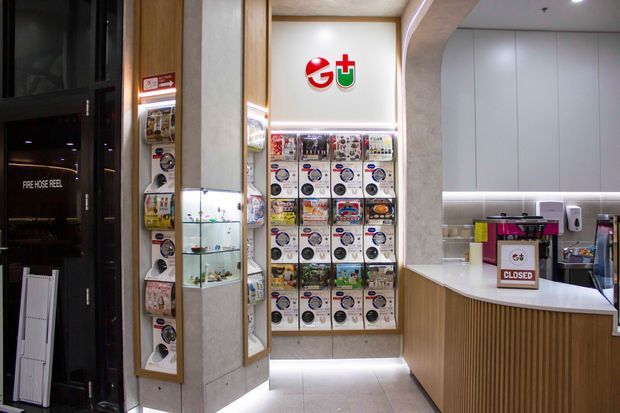 A compact, contemporary retail interior with a brightly illuminated gachapon capsule-toy display wall, glass display shelving, and a reception or service counter. The fit-out uses pale stone-look wall surfaces, warm vertical timber panelling, white cabinetry, integrated LED strip lighting, and dark-framed glazing. Product displays and branded signage dominate the space.