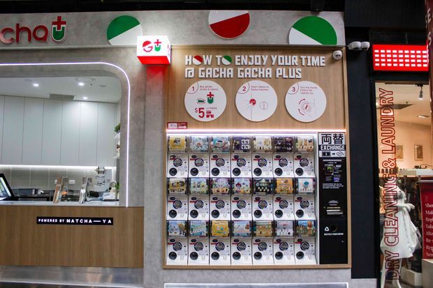 A compact commercial gacha capsule-toy shopfront inside an indoor retail setting. The frontage features a wood-toned service counter, white built-in cabinetry, grey wall panels, illuminated signage, a large grid of capsule vending machines, and instructional graphics. A neighbouring cleaning and laundry storefront is partially visible at the right.