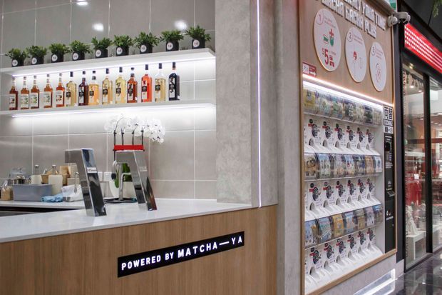 A compact, contemporary commercial beverage and dessert kiosk or café counter. The space features a white countertop, wood-look front cabinetry, illuminated shelves displaying bottled drinks, potted plants, coffee or beverage equipment, and a branded product display wall beside a glass-front refrigerator. The palette is light and neutral with warm timber accents and integrated LED lighting.