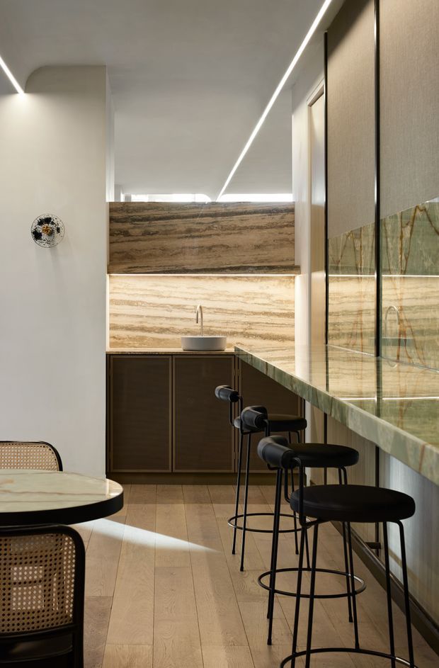 A refined contemporary kitchen and dining area with a compact peninsula or breakfast bar, dark timber cabinetry, a small vessel basin, and dramatic horizontally veined stone surfaces. A full-height glazed partition or reflective wall runs along the right side, while slim linear LED lighting is integrated into the ceiling. Pale timber flooring, black bar stools, and a rounded dining table with cane seating create a warm, minimalist interior.