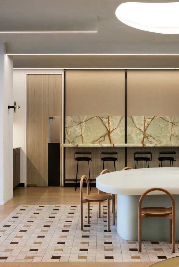 A refined contemporary hospitality-style interior featuring a large rounded white island or dining counter in the foreground, curved timber chairs, and a linear bar with black stools along the rear wall. The space combines warm timber cabinetry and doors, a pale green veined stone backsplash, and a patterned light tile floor with small brown and dark inset squares. Concealed linear lighting and a large oval ceiling light create a softly illuminated, sophisticated atmosphere.