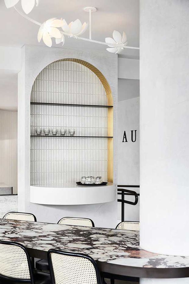 A refined dining area featuring a large rounded marble dining table with a dark edge, woven cane dining chairs with black frames, and a sculptural built-in arched display niche. The niche has a vertically ribbed light wall finish, gold-toned curved trim, black glass shelves, and small glassware. White sculptural floral pendant lights hang overhead, while black railing details and minimalist white walls complete the contemporary setting.