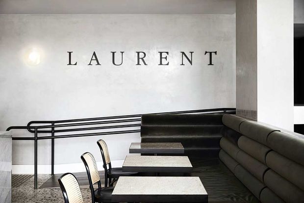 A refined restaurant or hospitality interior with a long upholstered banquette, small marble-topped tables, black-and-white dining chairs, and black metal railings. The pale textured wall features large “LAURENT” lettering, while dark flooring and monochrome furnishings create a sophisticated, graphic atmosphere.