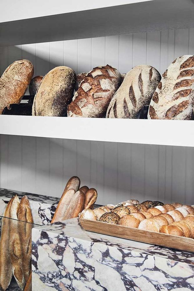 Close-up view of a stylish bakery display featuring artisan loaves arranged on white open shelving and in a wooden bread tray. The display is set against vertically panelled white walls, with a dramatic veined marble counter in white, grey, and dark purple tones in the foreground.