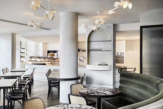 A refined open-plan hospitality or residential entertaining space with a dining area in the foreground, a curved upholstered banquette, marble-topped round tables, and woven-back chairs. The bright kitchen or service area is visible beyond, along with a sculptural arched display niche, ribbed glass door, concrete cylindrical column, and distinctive botanical pendant lights.