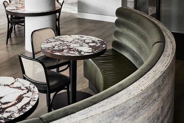 A refined contemporary dining or hospitality interior featuring a dramatic curved upholstered banquette, small round marble-topped tables with dark veining, and black cane-back dining chairs. The space has dark timber flooring, pale walls, and a sculptural, high-end material palette.