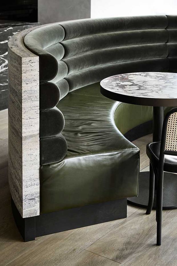 Close-up interior view of a dramatic curved banquette upholstered in dark olive-green leather, featuring vertically segmented rounded back cushions and a pale textured stone side panel. A round marble-topped dining table with a dark base and black dining chair sit in front, over a warm timber-look floor. The composition is refined and luxurious with strong sculptural detailing.