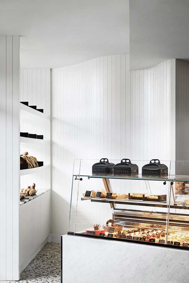 Bright, minimalist commercial food retail interior, likely a bakery or patisserie. The space features white vertically panelled walls, integrated recessed display niches, terrazzo flooring, and a glass refrigerated display counter with a pale stone-faced base. Black product bags and neatly arranged pastries provide restrained contrast.