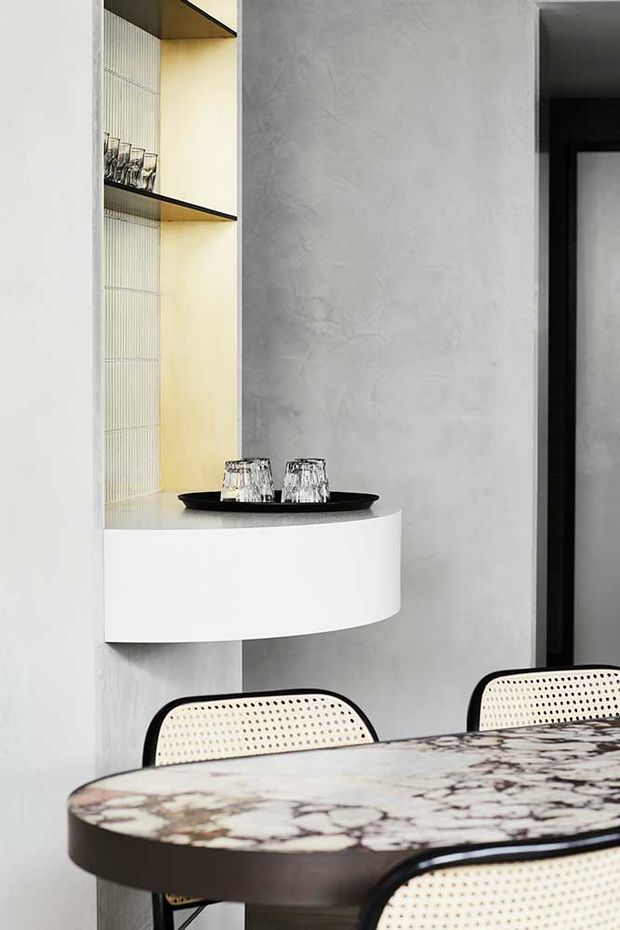 A refined dining area featuring a round or oval marble-patterned dining table with a dark edge, woven cane dining chairs, and a curved white floating console or bar surface. Behind it is a built-in open shelving niche with warm backlighting and glassware, set against a textured pale grey wall. A dark-framed doorway is visible to the right.