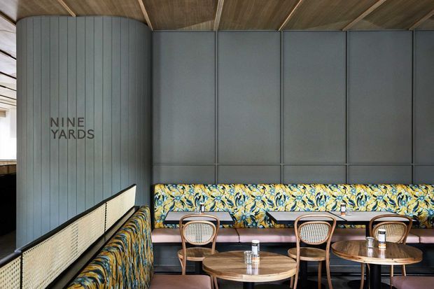 A stylish restaurant or hospitality dining area with a long upholstered banquette, round wooden tables, cane-back chairs, dark grey wall panelling, and a richly patterned botanical fabric accent. The ceiling uses warm timber boards with exposed beams, while woven screen panels and curved wall forms add texture and visual interest. The “Nine Yards” signage identifies the venue.
