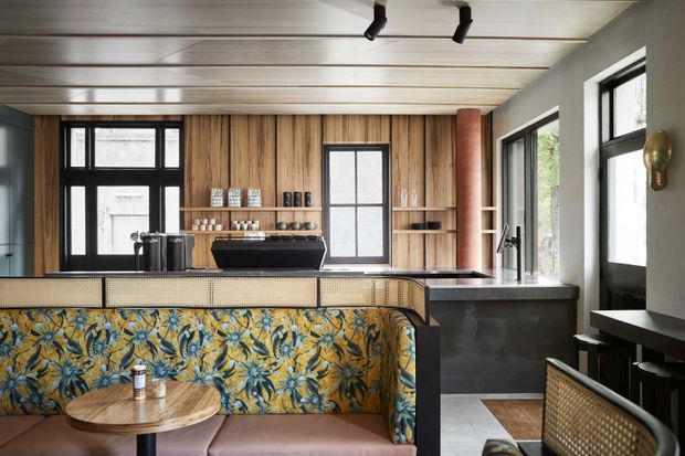 A stylish open-plan kitchen and dining nook featuring warm timber wall and ceiling panelling, black-framed windows, dark stone kitchen surfaces, and a patterned upholstered banquette with a round timber table. Open shelving and integrated cabinetry add functional storage, while track lighting and a sculptural wall light complete the space.