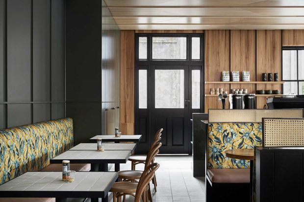 A stylish café or restaurant interior with a row of tiled dining tables and curved wooden chairs, patterned upholstered banquette seating, a dark service counter, and open shelving displaying cups and coffee equipment. The space features warm timber wall and ceiling lining, black-framed glazed doors and windows, dark cabinetry, and a bold botanical-patterned upholstery scheme.