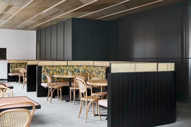 A contemporary restaurant or café interior with a long banquette seating area, pale timber and cane chairs, patterned upholstered bench seating, and dark vertical-panel cabinetry or wall cladding. The ceiling features warm timber slats, while the floor and surrounding surfaces are light and understated.