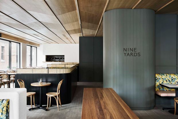 A refined hospitality-style interior with a bar and dining area. The space features dark cabinetry, a curved vertically panelled feature wall with “Nine Yards” signage, a warm timber-lined ceiling, large windows, stone-topped counters, patterned banquette seating, and timber tables. The palette combines dark charcoal, warm wood, muted grey, and colourful upholstery accents.
