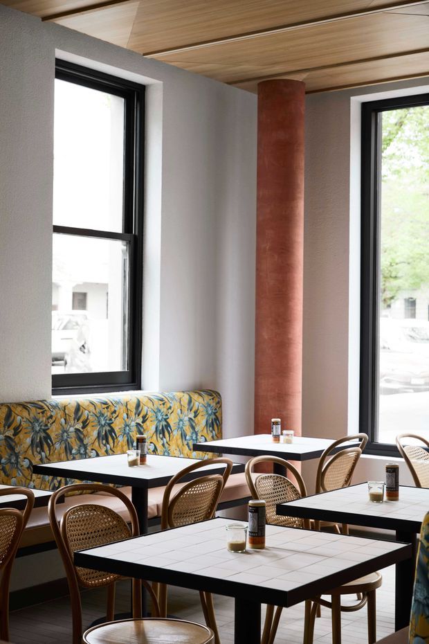 A warmly lit contemporary café or restaurant interior with multiple white tiled square tables, bentwood and cane dining chairs, and a patterned upholstered banquette along the wall. Tall black-framed windows bring in daylight, while a cylindrical terracotta-toned column and linear timber ceiling panels add strong architectural features. The space has a refined, slightly retro character with botanical-patterned seating and neutral plaster walls.