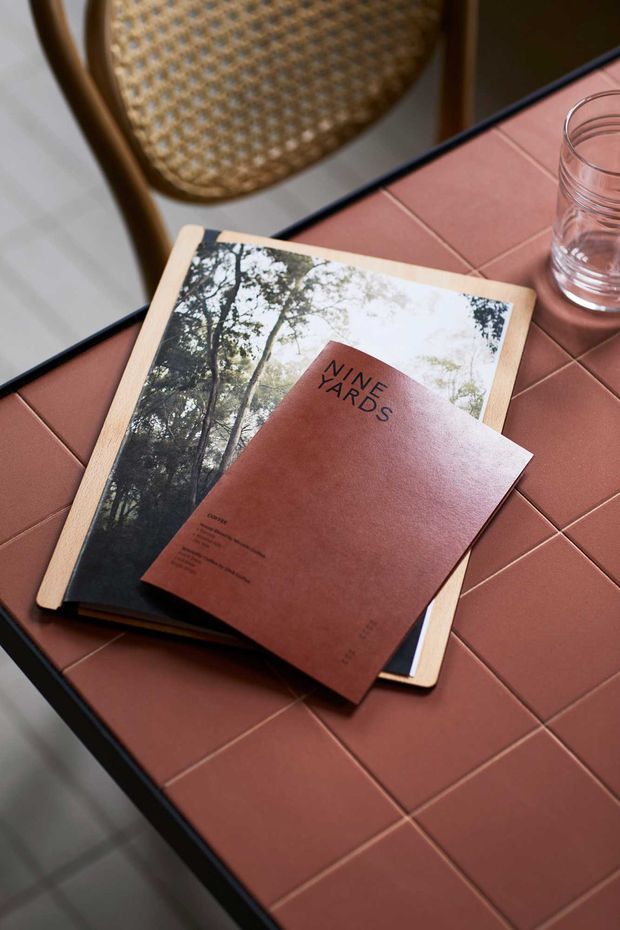 Close-up styled interior detail showing a terracotta-toned tiled tabletop with dark edging, a woven timber-framed chair, a clear drinking glass, and stacked books or magazines. The scene has a warm, tactile, editorial feel with earthy colours and natural textures.