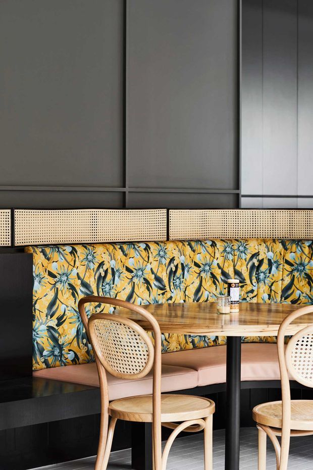 A close-up of a stylish restaurant or hospitality dining booth. The space features a bold yellow and blue floral upholstered banquette, pale pink seat cushions, cane-backed bentwood chairs, a round timber table with a black pedestal base, dark built-in joinery, a perforated cane screen, and large grey wall panels. The composition is warm, graphic, and eclectic with strong contrast between the floral upholstery and dark architectural surfaces.