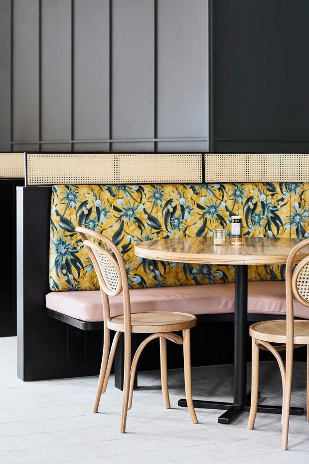 A stylish dining or café booth area with a long upholstered banquette, bentwood cane chairs, and a round timber table on a black pedestal base. The booth features a vivid botanical floral fabric in yellow, blue, and green, with cane detailing along the upper backrest. Dark vertical wall panelling and black built-in joinery create a strong backdrop, while pale tiled flooring brightens the space.