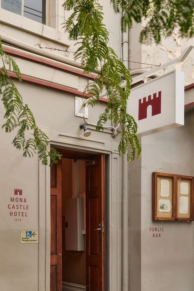 Exterior view of the Mona Castle Hotel public bar entrance. The narrow heritage-style facade has pale rendered walls, dark red trim, timber panelled entry doors, exposed service conduits, climbing greenery, wall-mounted signage, a projecting hotel sign, and framed notices beside the doorway.
