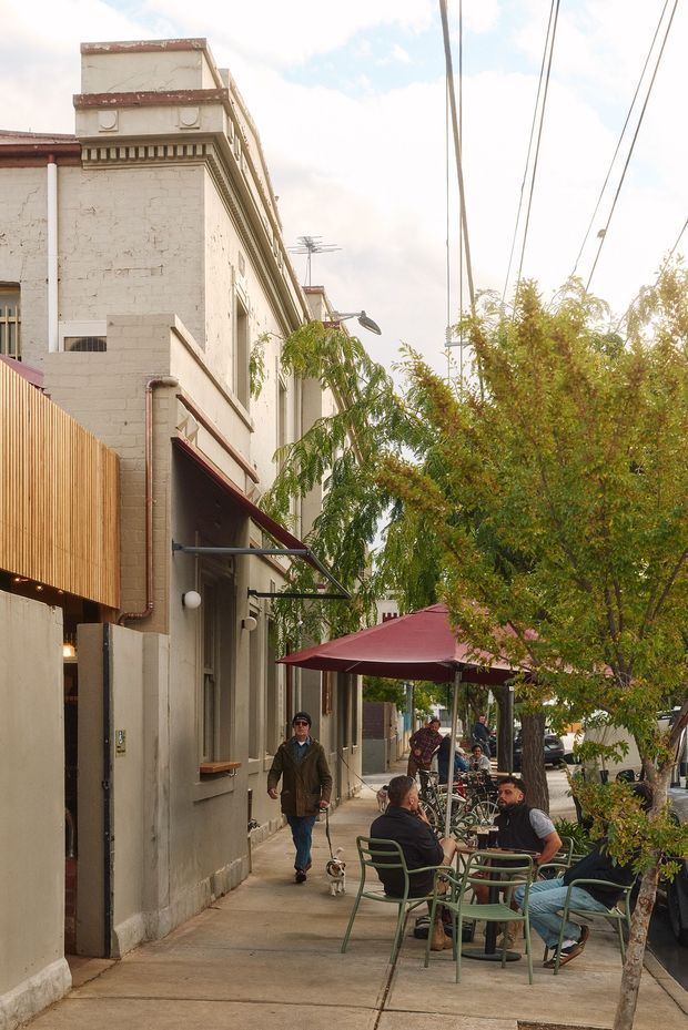 Street-level view of a neighbourhood café or commercial frontage on a tree-lined urban sidewalk. The pale masonry building has decorative classical cornice detailing, projecting awnings, exposed utility wiring, and outdoor seating beneath burgundy umbrellas. People, bicycles, and small trees occupy the footpath dining area.