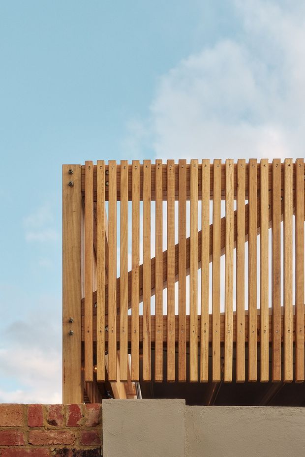 Close-up exterior architectural detail of a timber slatted balcony or screening structure. Vertical warm-toned wooden battens are fixed to a larger timber frame, with a diagonal timber stair or structural element visible behind them. The structure sits above a rendered wall and an older brick boundary wall, against an open blue sky. The image emphasizes joinery, rhythm, texture, and timber construction rather than an identifiable interior room.
