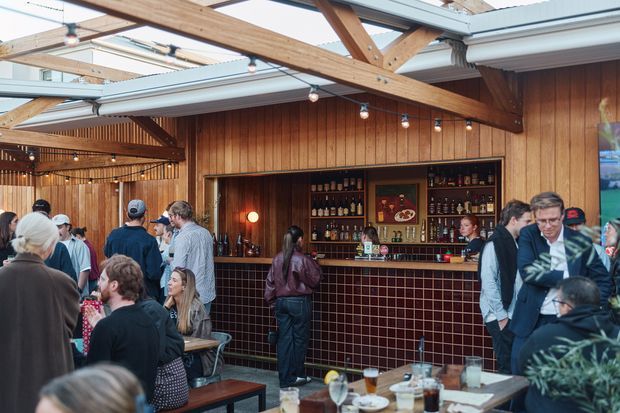 A busy, warmly lit bar and dining area in a timber-framed hospitality venue. The space features extensive vertical timber wall panelling, exposed heavy timber roof beams, clerestory-style glazing and an open roof structure. A built-in bar with dark cabinetry, bottle shelving and a burgundy-red tiled front dominates the rear wall, while patrons gather around timber tables and benches. Festoon-style pendant bulbs and warm lighting create an inviting atmosphere.