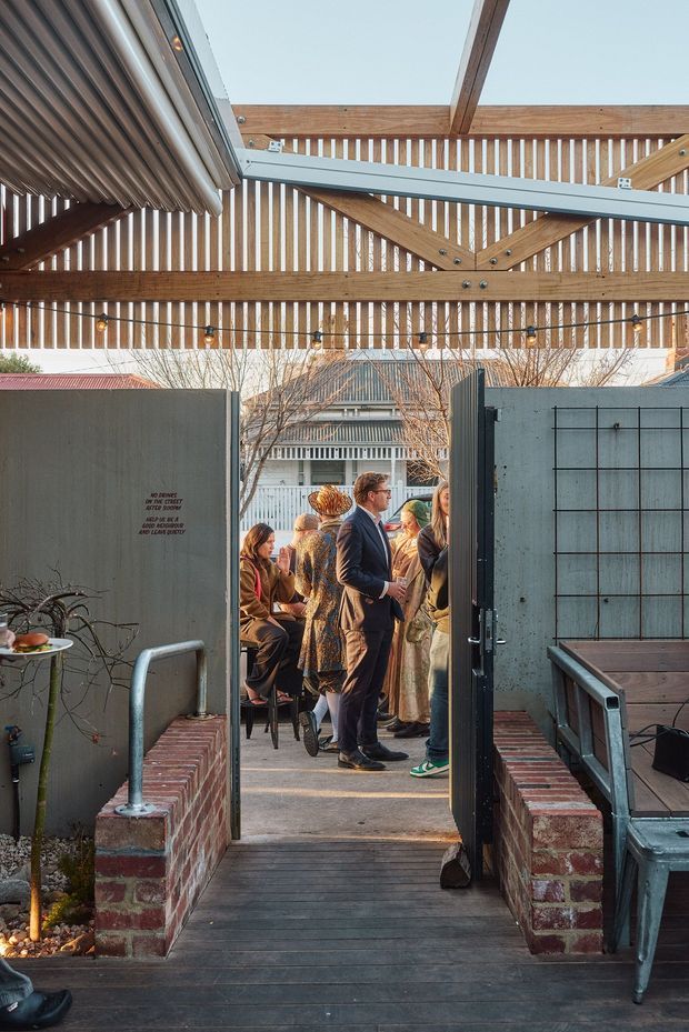 A covered outdoor terrace or courtyard entrance with a timber slatted overhead structure and warm string lights. A black metal gate opens toward a gathering of people, while brick planter walls, dark timber decking, concrete privacy panels, and metal outdoor seating define the space. The image strongly emphasizes the architectural canopy, entry threshold, and semi-enclosed outdoor hospitality setting.