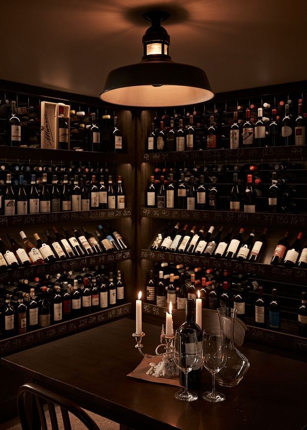 A dark, intimate wine storage room lined with extensive built-in shelving filled with wine bottles. Chalkboard-style shelf labels, dark wood cabinetry, a central pendant light, and a tasting table with wine glasses, a decanter, candles, and chairs create a moody cellar atmosphere.
