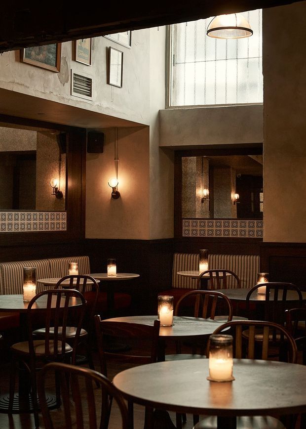 Atmospheric restaurant or café dining interior with multiple round tables, dark timber bentwood chairs, striped banquette seating, candlelit table lamps, decorative patterned tile dado panels, framed artwork, wall sconces, and large industrial-style glazing. The space has a moody, warm, vintage-inspired character with textured plaster walls and dark timber detailing.