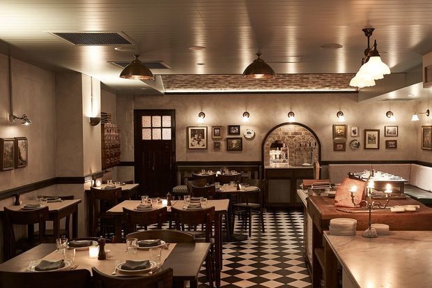 A warmly lit, richly detailed restaurant dining room with dark timber tables and chairs, set tables, framed artwork, pendant lighting, a patterned black-and-white tiled floor, and a curved brick-lined service opening. A timber-topped bar or service counter occupies the right foreground.