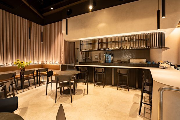 A warmly lit contemporary hospitality-style interior featuring a bar and dining area. The space has a long dark timber bar with stools, a curved pale stone or solid-surface counter, small dining tables and black chairs, a tiled backsplash, and sculptural vertical ribbed wall and ceiling detailing. Concealed and pendant lighting create an intimate, refined atmosphere.