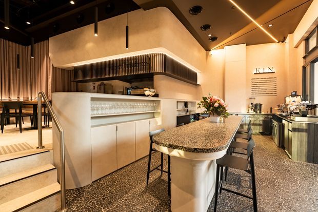 A warm, sophisticated hospitality-style interior featuring a bar and café counter with a large textured stone island, integrated display shelving, commercial coffee equipment, and bar stools. A raised dining area with tables and chairs is visible at left, accessed by a short stair. The space has dark ceilings, curved illuminated bulkheads, linear pendant lighting, timber slat detailing, and polished flooring.