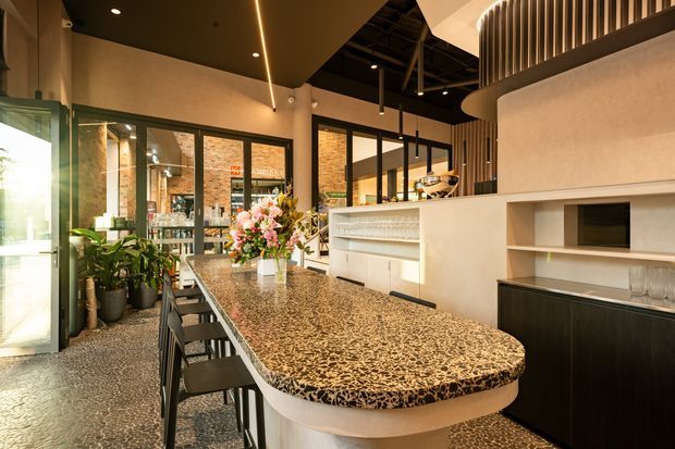 Contemporary hospitality-style interior featuring a long curved bar island with a speckled stone benchtop, integrated shelving, dark cabinetry, black-framed glazing, exposed ceiling services, and vertical slatted detailing. Plants and floral arrangements add warmth to the refined space.