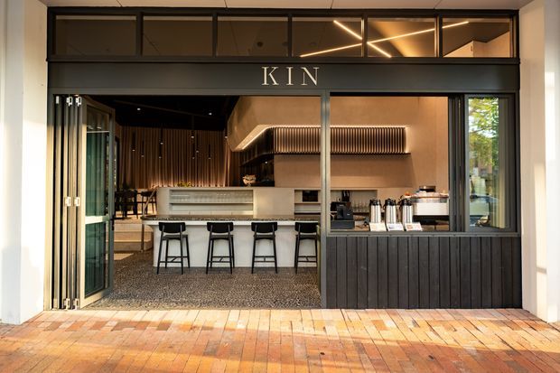Street-facing view into a contemporary hospitality venue branded “KIN,” with a dark framed glazed façade, folding glass doors, timber-clad lower panels, a bar counter with stools, warm timber interiors, and distinctive linear pendant lighting. The foreground is a brick-paved exterior walkway.