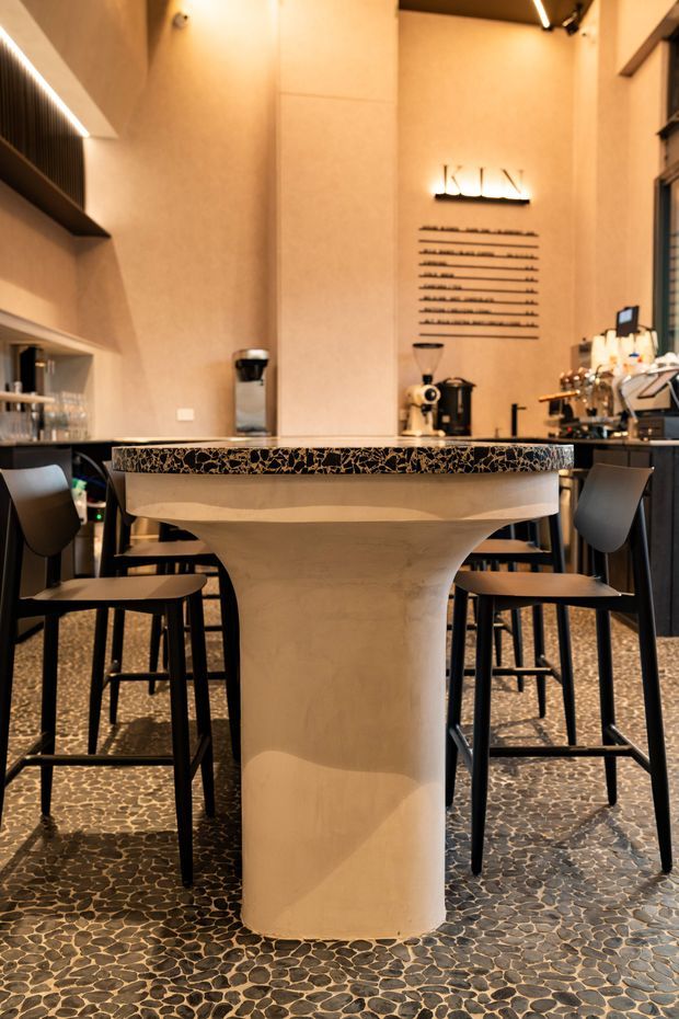 A warmly lit contemporary café interior focused on a sculptural central bar table with a black-and-tan patterned stone or terrazzo top and a curved beige pedestal base. Black bar stools surround the table, while a coffee service counter, espresso equipment, shelving, and a wall-mounted “KLIN” sign are visible behind it. The floor is finished with small irregular dark stone or pebble-like tiles, and linear integrated lighting adds a refined atmosphere.