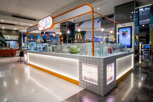 A brightly lit contemporary juice and dessert kiosk in a shopping-centre food court, with a tiled service counter, illuminated white ribbed panels, orange tubular framing, pendant lights, digital menu screens, glass display vessels, and visible surrounding food outlets.