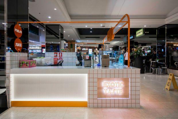 A contemporary shopping-centre food and beverage kiosk with a tiled service counter, illuminated ribbed front panel, orange accents and overhead orange framing. Dark glossy shopfronts, seating, mall circulation space and illuminated signage are visible in the background.