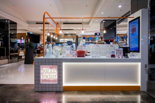 A contemporary retail juice and fresh-fruit kiosk in a shopping-centre setting. The compact counter features a tiled front panel with illuminated “REAL FRUIT” signage, a ribbed translucent front detail with warm underlighting, glass beverage dispensers, display refrigerators, pendant lighting, and bright digital menu screens. The surrounding mall interior is visible in the background.
