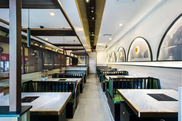 Interior view of a contemporary restaurant or dining venue with rows of booth seating and square dining tables. The space features black metal framing, dark timber accents, pale wall panelling, polished light flooring, arched wall niches with artwork, and linear recessed lighting. The layout is long and narrow with a modern commercial fit-out.