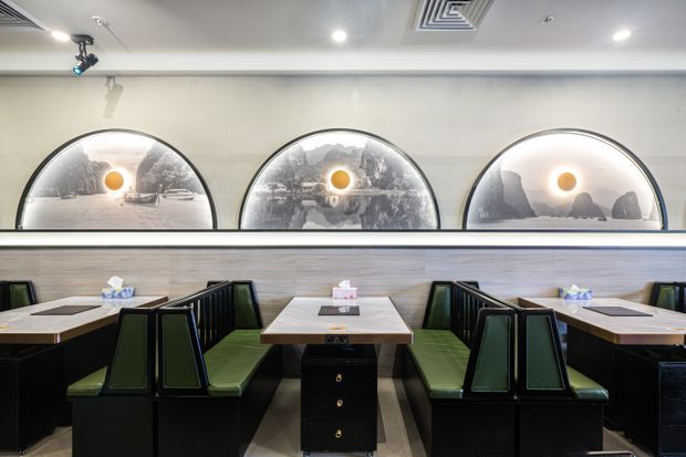A contemporary commercial dining interior with rows of booth seating arranged along a feature wall. The booths have dark framing and deep green upholstered seats, paired with light rectangular tables and central black storage pedestals. A horizontal wood-look wall panel runs beneath three large arched illuminated decorative panels, while recessed ceiling lighting and exposed air-conditioning vents complete the space.